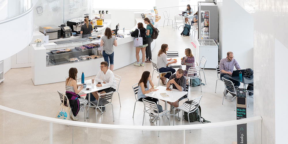 People are sitting at tables in the Biozentrum cafeteria, eating, drinking and talking. In the background, some people stand at the serving counter.